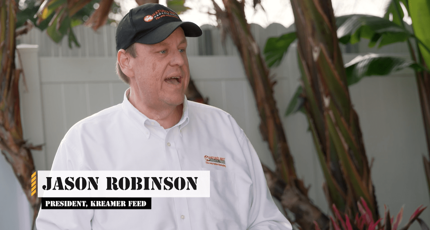 Business leader in white button-down with Nature’s Best Organic Feeds logo, addressing camera in a garden.