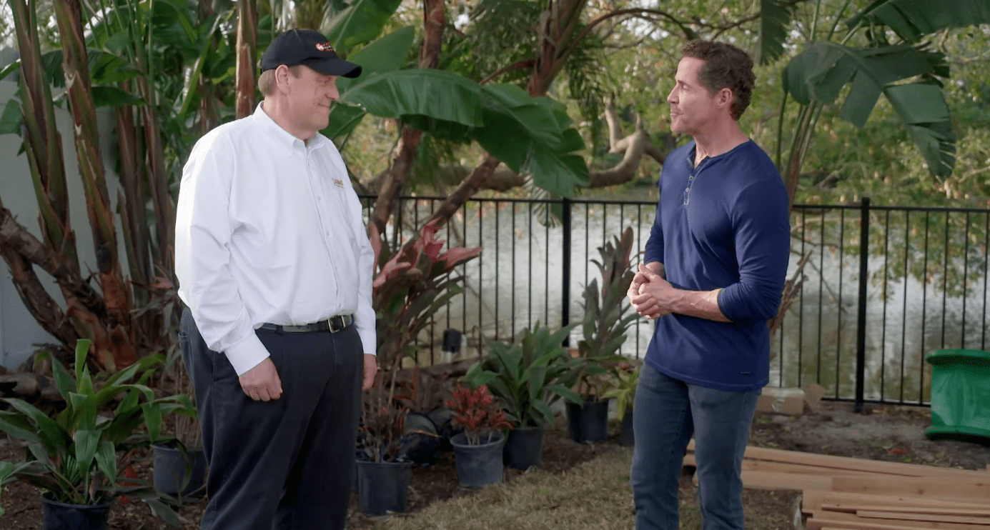 Two people discussing a backyard project beside potted plants and a fence; Organic Feeds shirt visible.